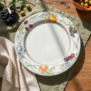 May include: White ceramic plate with a decorative border featuring illustrations of fruits like pears, blackberries, and cherries. The plate has a woven-style brown border and is set on a green cloth.