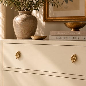 May include: A cream-colored dresser featuring two gold-toned metal drawer pulls shaped like leaves. The dresser top holds a large speckled ceramic vase with green branches, two small brass bowls, and two stacked books titled Houses Atelier AM and Live Beautifully.