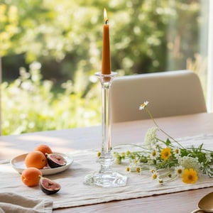 May include: A tall, clear glass candlestick holder with a hexagonal base and a lit, slender, golden-yellow taper candle. The candlestick is positioned on a light-colored fabric runner next to a small plate of fresh apricots and sliced figs.