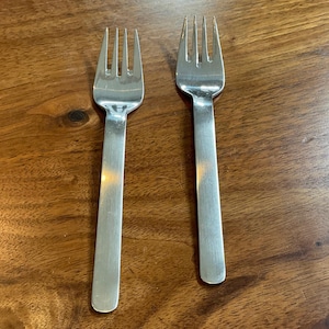 May include: Two stainless steel forks with four tines each, lying on a wooden surface. The forks have a simple, modern design with long, rectangular handles. The polished metal reflects the light, creating highlights.