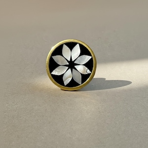May include: A circular knob featuring a gold-toned metal rim and a black center inlay. The inlay displays a white, iridescent mother-of-pearl floral pattern composed of eight petal-shaped segments arranged in a symmetrical star-like design.