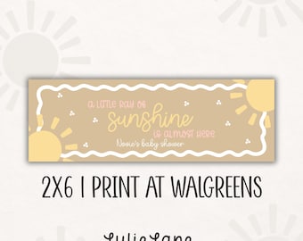 May include: A rectangular tan sign with a white wavy border featuring yellow sun illustrations and the text A little ray of sunshine is almost here Novie's baby shower. The sign includes the text 2x6 I print at Walgreens and Lulie Lane.