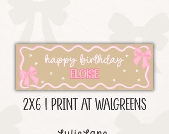 May include: A rectangular tan banner featuring a pink scalloped border, two pink bows, and white polka dots. The text reads "happy birthday ELOISE" in white and pink. Below the banner, the text states "2X6 | PRINT AT WALGREENS" and "Lulie Lane".