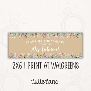 May include: A rectangular beige sign with floral borders and the text "FRESH OFF THE MARKET THE FUTURE Mrs. Belmont". Below the text is "2X6 I PRINT AT WALGREENS" and the brand name "Lulie Lane".