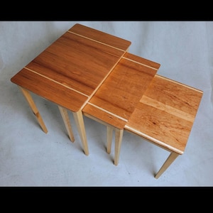 May include: A set of three nesting tables. Each table has a rectangular top with a light wood inlay and tapered legs. The wood has a warm, reddish-brown tone. The tables are arranged in a nested configuration.