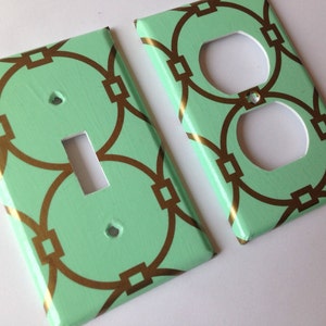 May include: Two decorative light switch and outlet covers with a mint green background and gold geometric pattern.