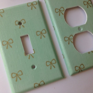 May include: Two light green wall plates with a gold bow pattern. One plate is for a light switch and the other is for an electrical outlet.