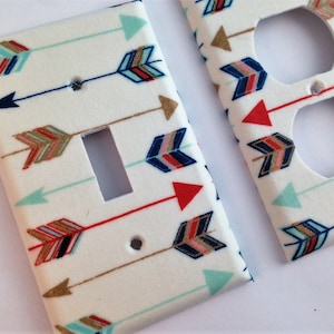 May include: Two white light switch and outlet covers with a pattern of colorful arrows. The arrows are in shades of red, blue, gold, and green.