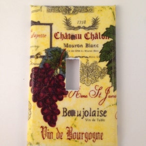 May include: A light yellow light switch cover with a vintage wine label design. The label features a bunch of grapes, the text "Château Chalon", "Mouron Blanc", "Vin de Bourgogne", and "Beaujolaise".