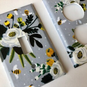 May include: Two light switch and outlet covers with a floral pattern. The covers are gray with white flowers, yellow centers, green leaves, and black bees. The pattern is repeated on both covers.