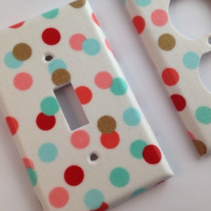 May include: Two white light switch and outlet covers with a polka dot pattern. The dots are red, pink, teal, and gold.