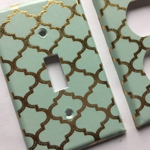 May include: A set of two light green and gold decorative wall plates. The plates feature a repeating geometric pattern with a gold metallic finish. One plate is for a light switch and the other is for an electrical outlet.