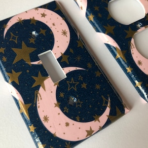 May include: Two decorative wall plates with a navy blue background and a pattern of pink crescent moons and gold stars. One plate is a light switch cover and the other is an outlet cover.
