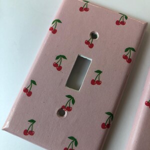 May include: Pink light switch cover with a repeating pattern of red cherries and green leaves. The cherries are painted on the cover, which has a rectangular shape with a single toggle switch opening and two screw holes.