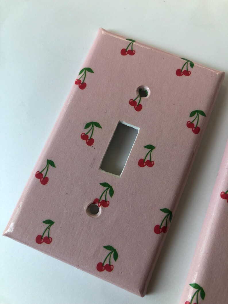 May include: Pink light switch cover with a repeating pattern of red cherries and green leaves. The cherries are painted on the cover, which has a rectangular shape with a single toggle switch opening and two screw holes.