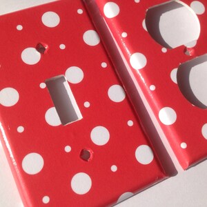 May include: Two red electrical outlet covers with a white polka dot pattern. One cover has a rectangular opening for a light switch, and the other has two circular openings for outlets. The covers are made of a glossy material.