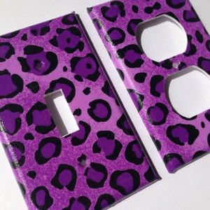 May include: Two purple and black leopard print light switch and outlet covers. The switch cover has a single toggle switch. The outlet cover has two standard outlets.