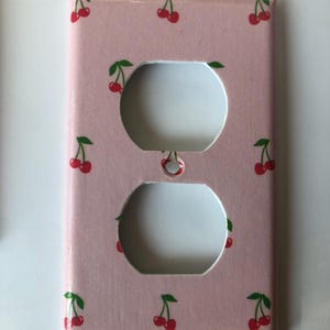 May include: A pink electrical outlet cover featuring a repeating pattern of red cherries with green stems and leaves. The cover has two circular openings for the outlets.