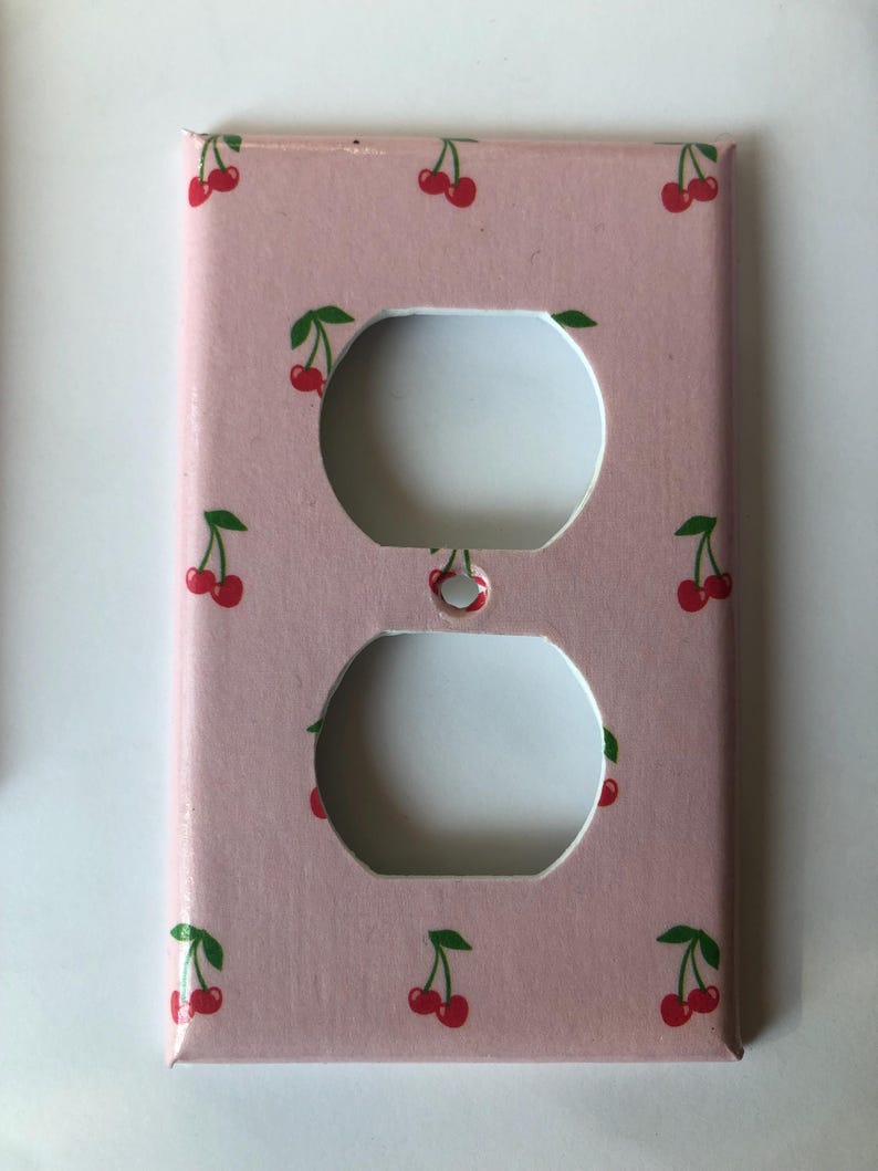 May include: A pink electrical outlet cover featuring a repeating pattern of red cherries with green stems and leaves. The cover has two circular openings for the outlets.