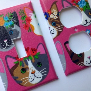 May include: Two pink light switch and outlet covers with a floral pattern featuring cats wearing flower crowns. The cats are grey, brown, and white.