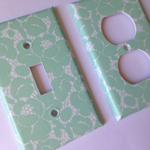 May include: Two light green wall plates with a white lace pattern. One plate is for a light switch and the other is for an electrical outlet.