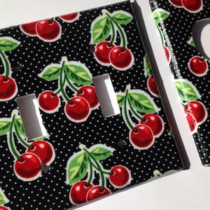 May include: Two black light switch and outlet covers with a red cherry pattern on a white polka dot background.