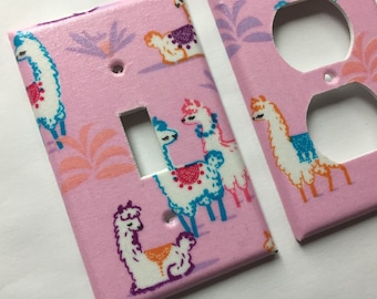 Pode incluir: Duas placas de interruptor rosa com um padrão repetido de lhamas coloridas e detalhes florais. Uma placa tem uma abertura para um interruptor, e a outra tem duas aberturas para tomadas. As lhamas são brancas com detalhes azuis, laranja e roxo.