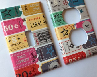May include: Two decorative light switch covers with a vintage ticket design. The covers feature colorful tickets with text like "Admit One," "Coupons," and "Family Fun." The color scheme includes pink, yellow, green, and gray.