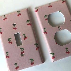 May include: Two decorative light switch covers. One has a single switch opening, the other has two electrical outlet openings. Both are painted light pink with a pattern of red cherries and green leaves.