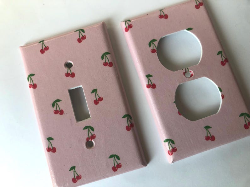 May include: Two decorative light switch covers. One has a single switch opening, the other has two electrical outlet openings. Both are painted light pink with a pattern of red cherries and green leaves.
