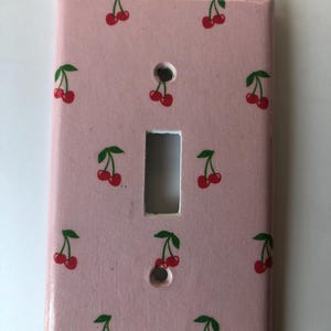 May include: A light pink light switch cover decorated with a pattern of red cherries and green leaves. The cover has two holes for screws and a rectangular opening for the switch.