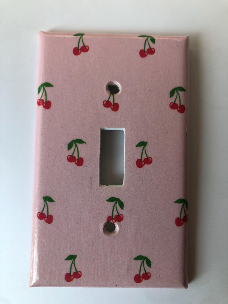 May include: A light pink light switch cover decorated with a pattern of red cherries and green leaves. The cover has two holes for screws and a rectangular opening for the switch.
