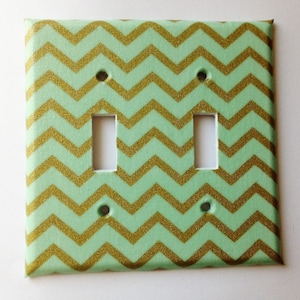 May include: A light green and gold chevron patterned light switch cover plate. The cover plate has two toggle switches.