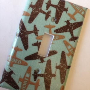 May include: A light blue light switch cover with a pattern of brown airplanes on it. The airplanes are in different sizes and orientations. The switch cover has a white light switch in the center.