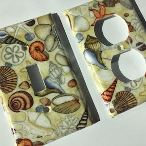 May include: Two light switch and outlet covers with a seashell design. The covers are white with a beige background and feature a variety of seashells, starfish, and other ocean-themed imagery.