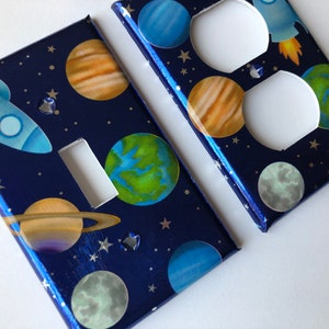 May include: Two decorative wall plates featuring a space theme. The plates are blue with silver glitter and feature planets, stars, and a rocket ship. One plate is a light switch cover and the other is a double outlet cover.