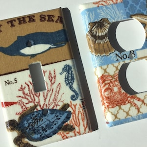 May include: Decorative light switch and outlet covers with a nautical theme. The covers feature marine life illustrations, including a whale, seahorse, and turtle, set against a beige, blue, and white patterned background. The text "BY THE SEA" is visible on one cover.