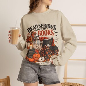 May include: A beige crewneck sweatshirt featuring a graphic of a skeleton reading a book in a red armchair. The text reads DEAD SERIOUS ABOUT MY BOOKS. The design includes pumpkins, sunflowers, a black cat, and a mug labeled GOOD BOOKS SPOOKY MOOD.