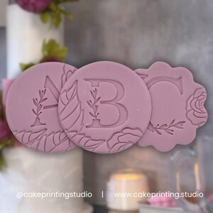 Può includere: Tre timbri rosa per torte con le lettere A, B e C. Ogni timbro presenta disegni floreali e di foglie attorno alle lettere. Il timbro con la lettera C ha un bordo smerlato. Lo sfondo include una torta e fiori.