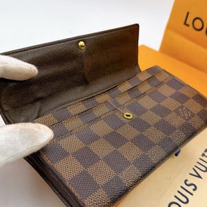 May include: Brown and tan checkered wallet with multiple card slots. The wallet is open, revealing the interior and a gold-colored snap closure. The wallet is made of a durable material and has a classic design. The brand name is visible on the packaging.