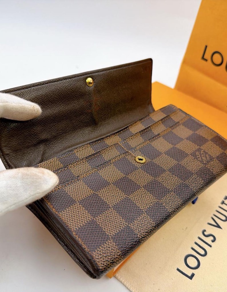 May include: Brown and tan checkered wallet with multiple card slots. The wallet is open, revealing the interior and a gold-colored snap closure. The wallet is made of a durable material and has a classic design. The brand name is visible on the packaging.