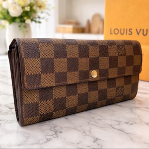 May include: Brown and tan checkered wallet with a gold-colored snap closure. The wallet is rectangular and has a flap closure. The background includes a shopping bag with the words "LOUIS VUI" printed on it.
