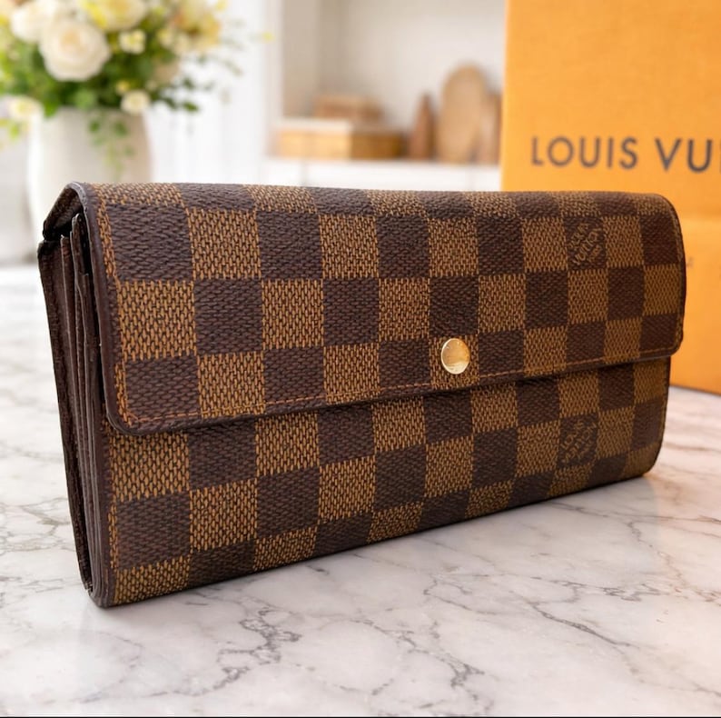 May include: Brown and tan checkered wallet with a gold-colored snap closure. The wallet is rectangular and has a flap closure. The background includes a shopping bag with the words "LOUIS VUI" printed on it.