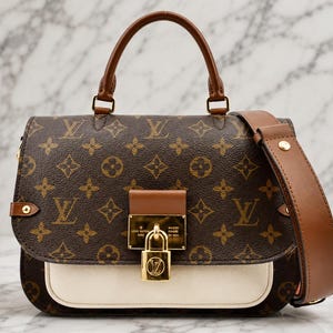 Op de afbeelding: Een bruine monogram handtas met een donkerbruin canvas met een herhalend logopatroon, een lichtbruin lederen handvat, een afneembare schouderriem, een crèmekleurig voorvak en een goudkleurig metalen hangslot met gegraveerde tekst.