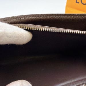 May include: A brown leather wallet with a silver zipper. The interior is a darker brown. The wallet is open, revealing the inside. The zipper is closed on the bottom and open on the top. The wallet is held by a person wearing a white glove.