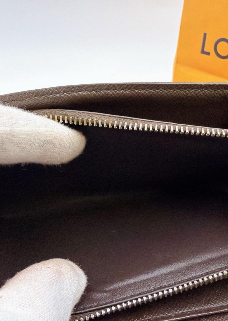 May include: A brown leather wallet with a silver zipper. The interior is a darker brown. The wallet is open, revealing the inside. The zipper is closed on the bottom and open on the top. The wallet is held by a person wearing a white glove.