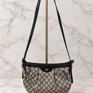 Puede incluir: Un bolso de hombro en forma de media luna con un patrón de monograma en beige y azul marino. El bolso tiene ribetes de cuero negro, cierre de cremallera superior y una correa larga de cuero negro. Un pequeño emblema dorado está centrado al frente.