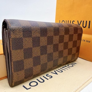 May include: Brown and tan checkered wallet. The wallet is rectangular and has a repeating pattern of brown and tan squares. The wallet is made of a leather-like material. The brand name "LOUIS VUITTON" is visible on the background.