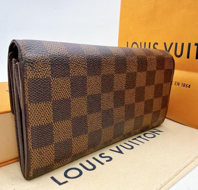 May include: Brown and tan checkered wallet. The wallet is rectangular and has a repeating pattern of brown and tan squares. The wallet is made of a leather-like material. The brand name "LOUIS VUITTON" is visible on the background.