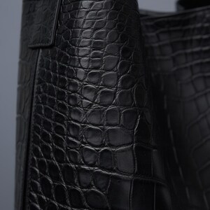 K&ouml;nnte beinhalten: Nahaufnahme einer schwarzen Lederhandtasche mit Krokodillederstruktur. Die Tasche hat ein strukturiertes Design mit einem Griff und einem Klappenverschluss. Das Leder scheint hochwertig zu sein und die Tasche ist wahrscheinlich ein Luxusartikel.